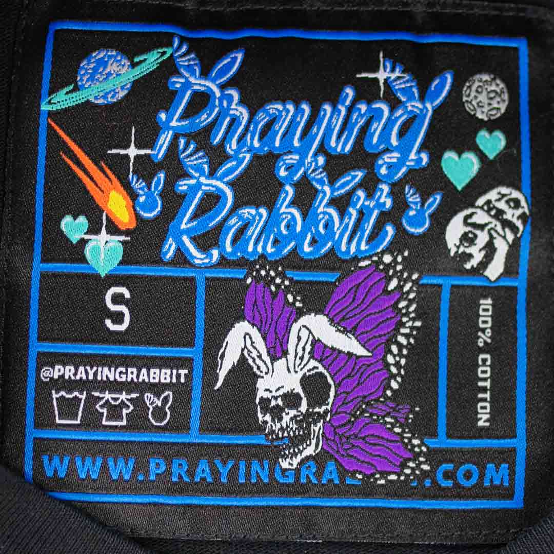 woven label that reads praying rabbit. it has a rabbit skeleton butterfly image with hearts, skulls, and comets. the planet saturn is also inside the tag