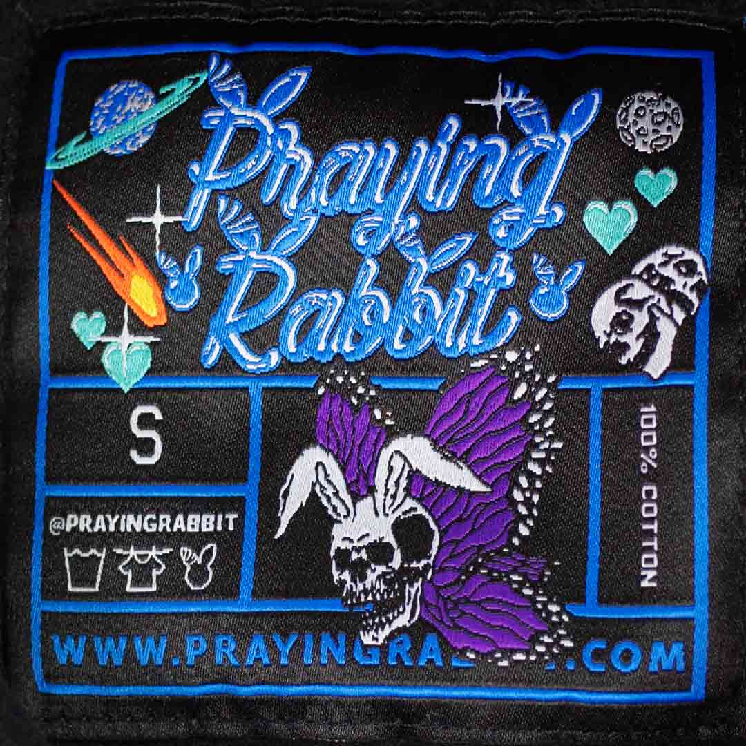 woven label that reads praying rabbit. the text is blue and in a nice cursive font. there is a rabbit skull butterfly, comets, skulls, hearts, and planets in the woven label