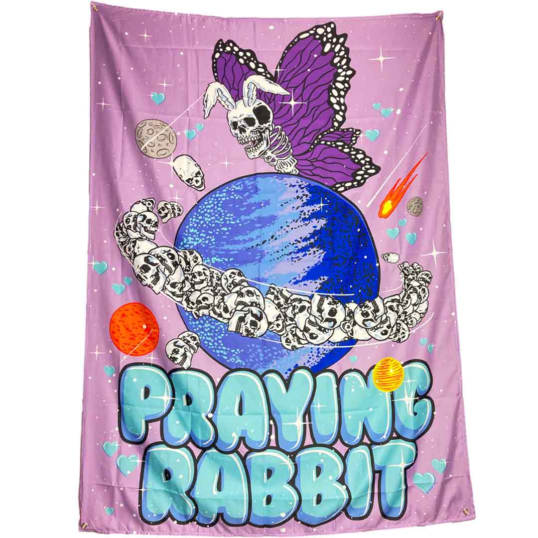 lavender tapestry with bubble letters that reads praying rabbit. above the letters is a graphic of a rabbit skeleton butterfly flying over the planet saturn. the planet saturn has skulls for rings. and there is hearts, sparkles, and comets around the entire design