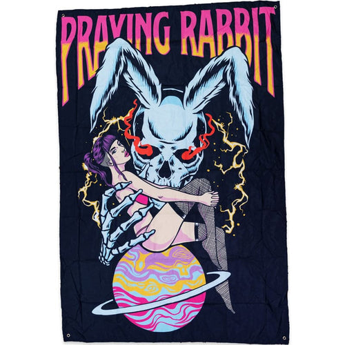 black tapestry with a graphic that shows a large blue rabbit skull holding a beautiful woman. the woman is sitting on the planet saturn and she is holding on to her knees. the blue rabbit skeleton has red glowing eyes. the girl has purple hair and fishnets on. there is text above the design that reads praying rabbit
