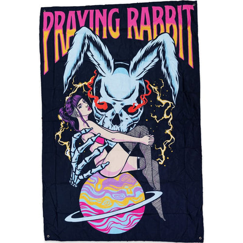 black tapestry with a graphic that shows a large blue rabbit skull holding a beautiful woman. the woman is sitting on the planet saturn and she is holding on to her knees. the blue rabbit skeleton has red glowing eyes. the girl has purple hair and fishnets on. there is text above the design that reads praying rabbit
